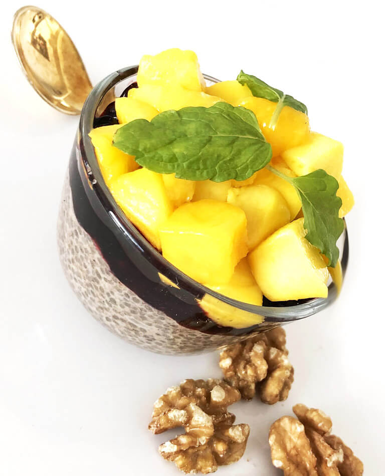 En skål med chiapudding med kokosmjölk toppad med mango och blåbär och valnötter vid sidan om.