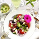 Ett fat med grekisk sallad med vegansk fetaost och avokado tzatziki serverat med gurkvatten och pyntat med en rosa blomma.