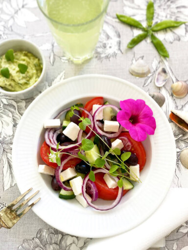 Ett fat med grekisk sallad med vegansk fetaost och avokado tzatziki serverat med gurkvatten och pyntat med en rosa blomma.
